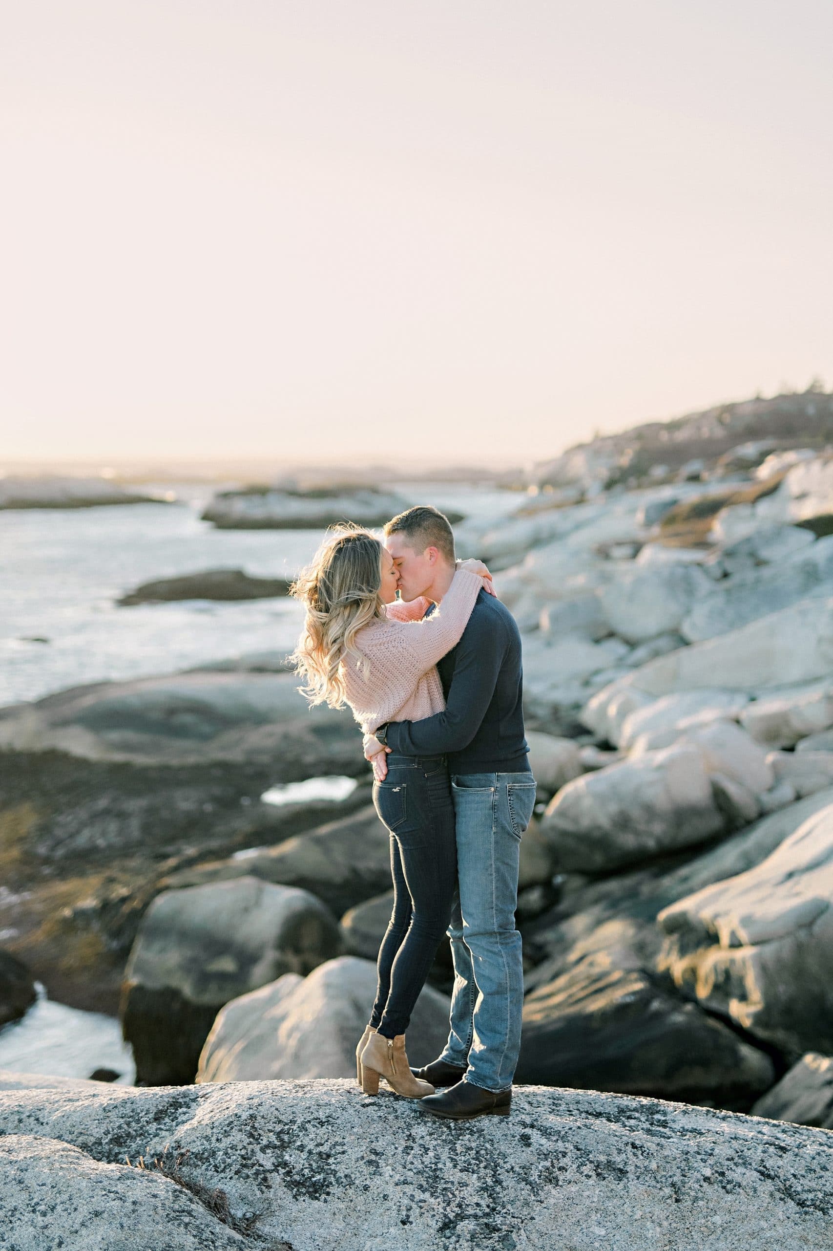 couple standing in embrace on rocks overlooking pink sky and ocean