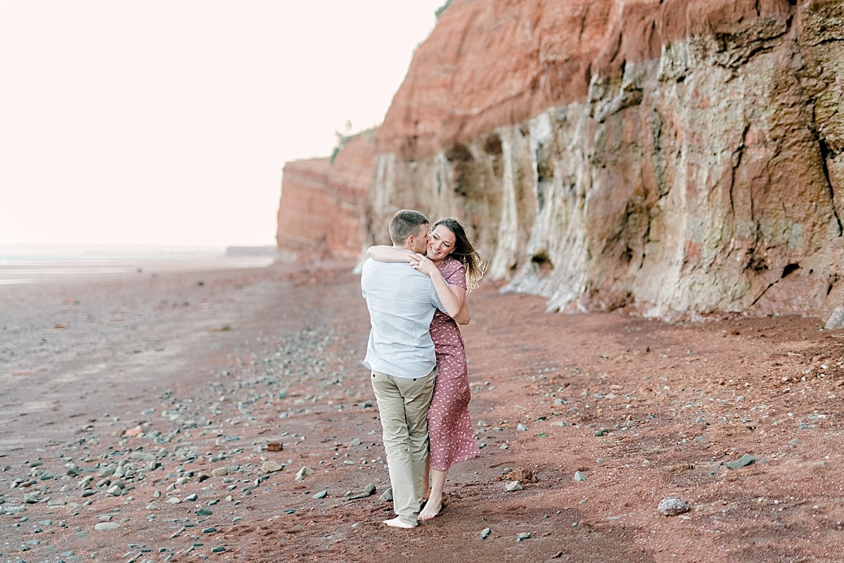 couple standing in front of giant tall red cliffs of blomidon beach during and engagement shoot