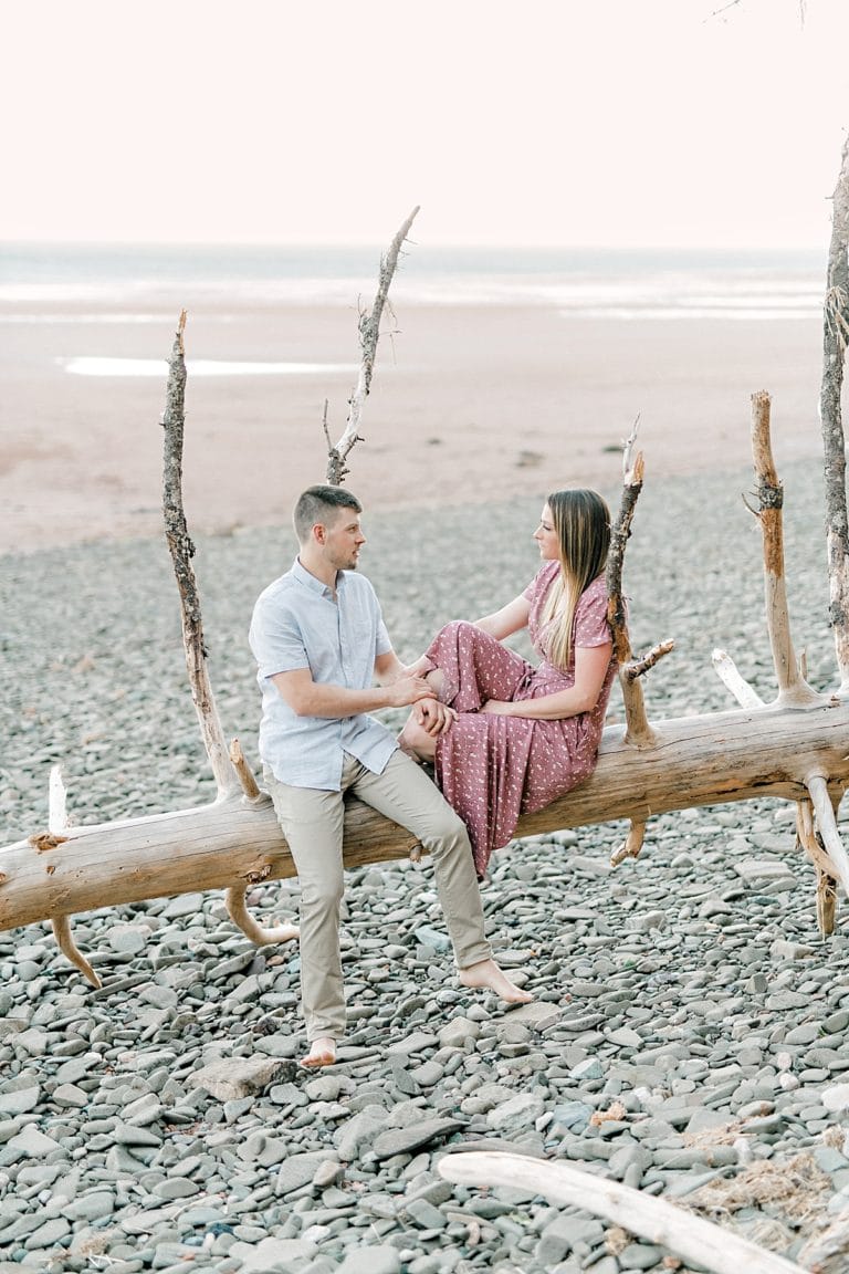 engaged couple sitting on a piece of driftwood that fell down on the beach at sunset