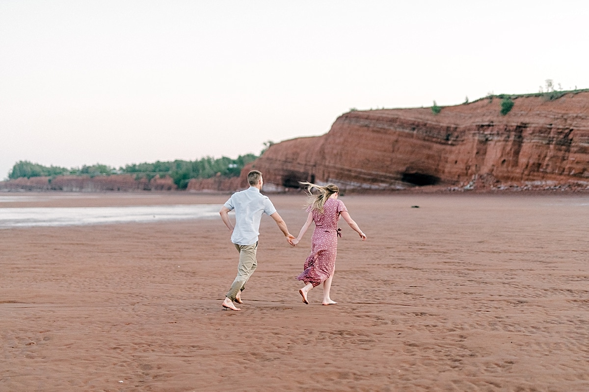 couple running in front of giant tall red cliffs of blomidon beach during and engagement shoot