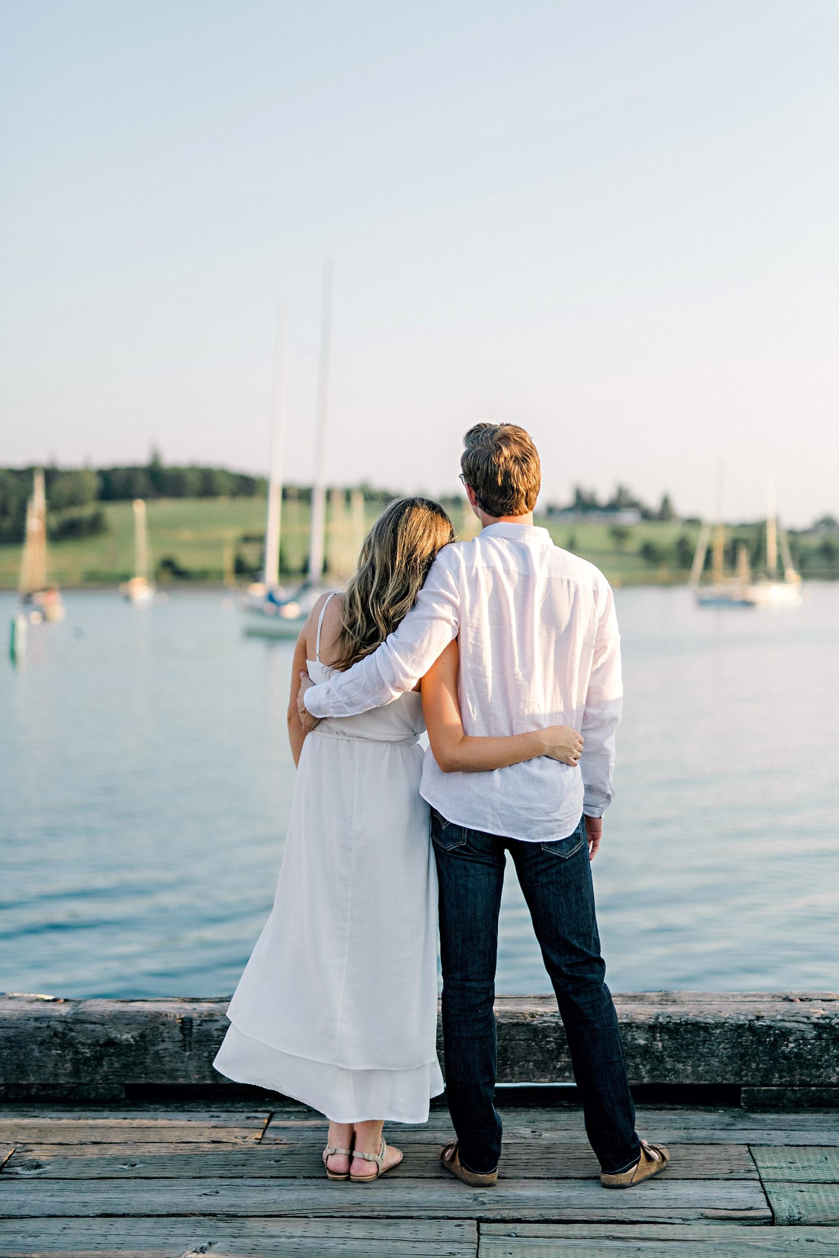 couple with their arms around eachother watching the sailboats on the ocean