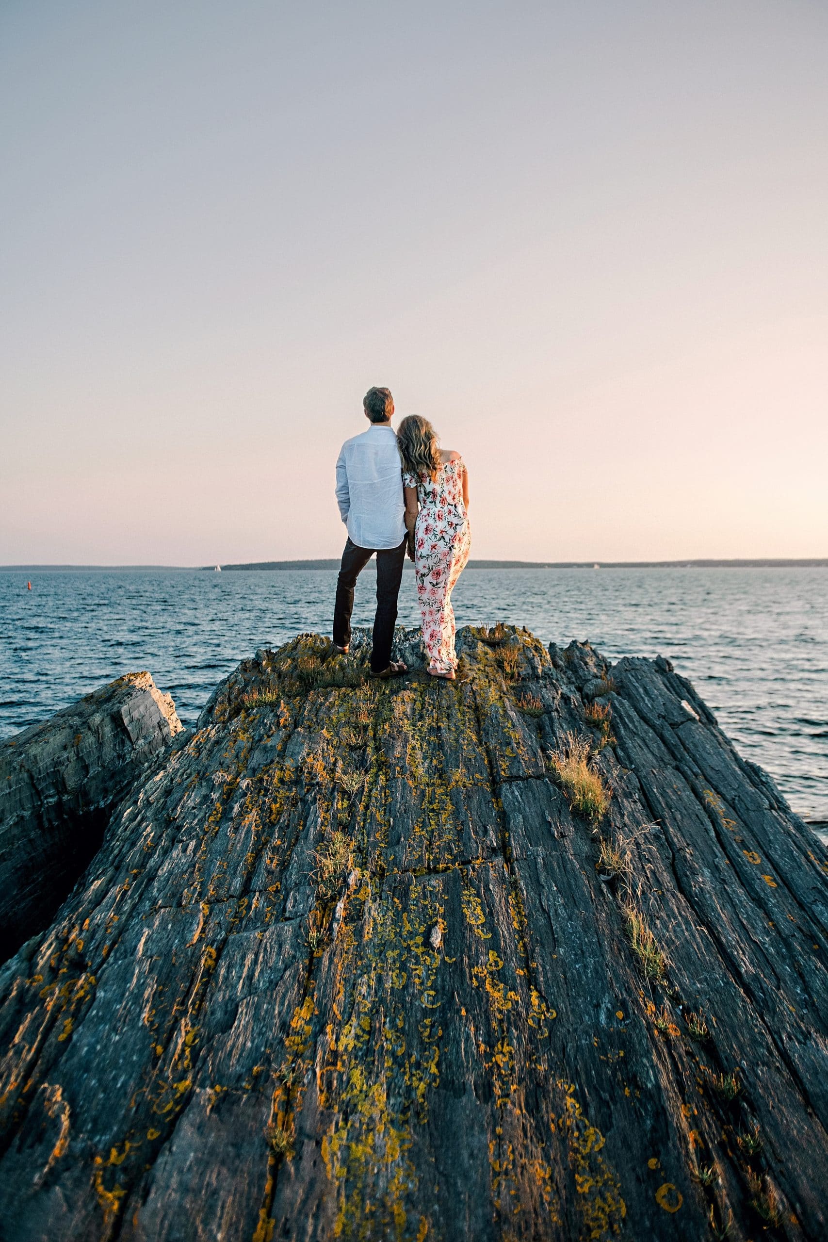 couple standing on top of rocky cliff over the ocean watching the sunset